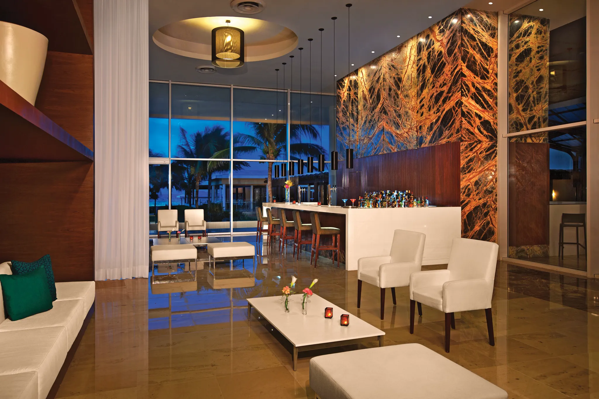 Moments lobby bar at Dreams Jade Resort & Spa featuring modern seating, illuminated bar, and ocean views through floor-to-ceiling windows.